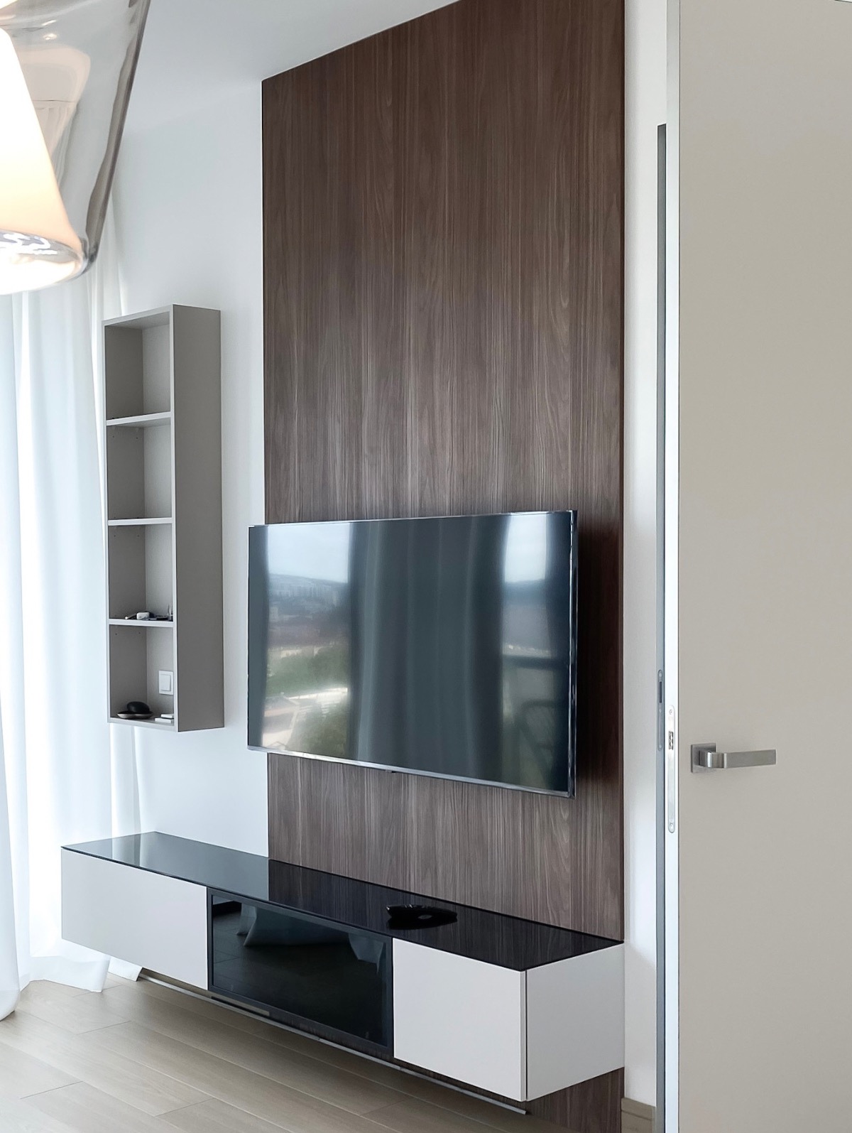 Nowoczesny salon z telewizorem na tle drewnianej ściany, szafka RTV z czarnym blatem, półka wisząca. Minimalistyczny design, jasne wnętrze, ukryte drzwi.