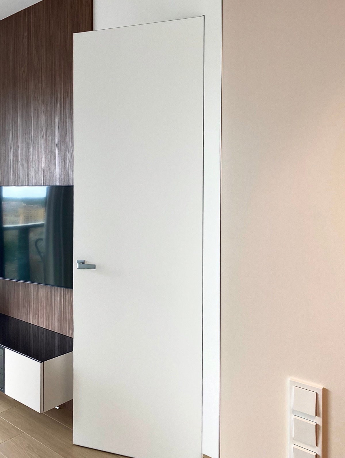 Minimalistyczne, białe drzwi wewnętrzne w nowoczesnym apartamencie w Szczecinie. Widoczny fragment ściany z telewizorem i drewnianą okładziną. Jasne, neutralne kolory.