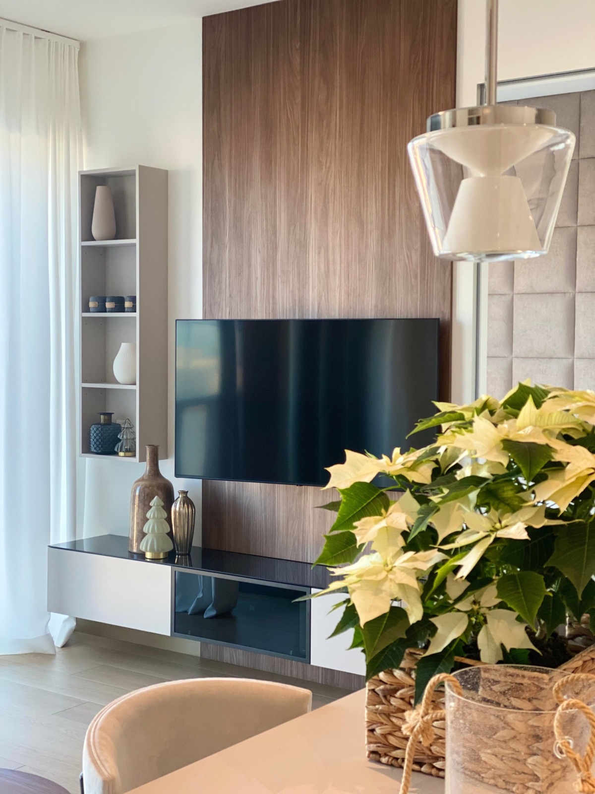 Nowoczesny salon z telewizorem na drewnianej ścianie, białą szafką RTV, półką z dekoracjami i lampą wiszącą. Wnętrze doświetlone naturalnym światłem.