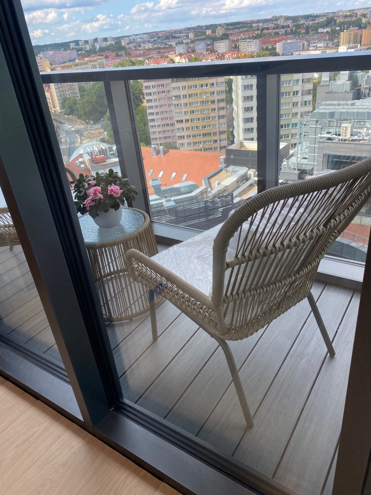 Nowoczesny balkon z rattanowym krzesłem i stolikiem z kwiatami, widok na miasto z wysokości. Szare deski podłogowe i czarne ramy okienne.