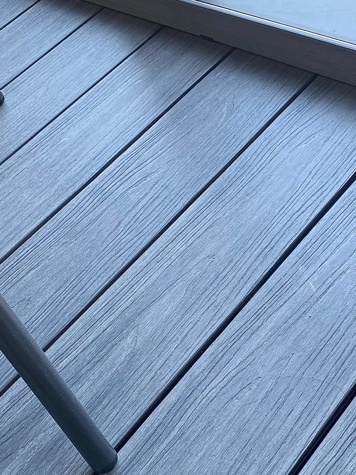 Szare deski tarasowe ułożone ukośnie, z widoczną nogą mebla w lewym dolnym rogu. Delikatna faktura drewna i regularne odstępy między deskami.