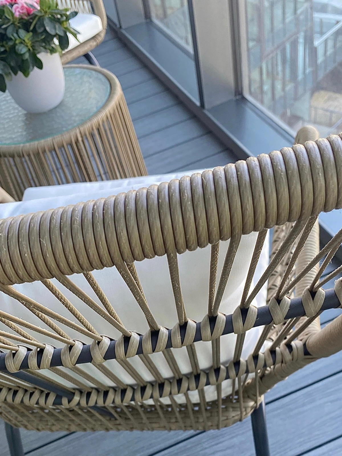 Detale rattanowego krzesła z jasną poduszką na balkonie z widokiem na miasto. Stolik z kwiatami w tle. Ujęcie z bliska, skupione na fakturze i wzorze.