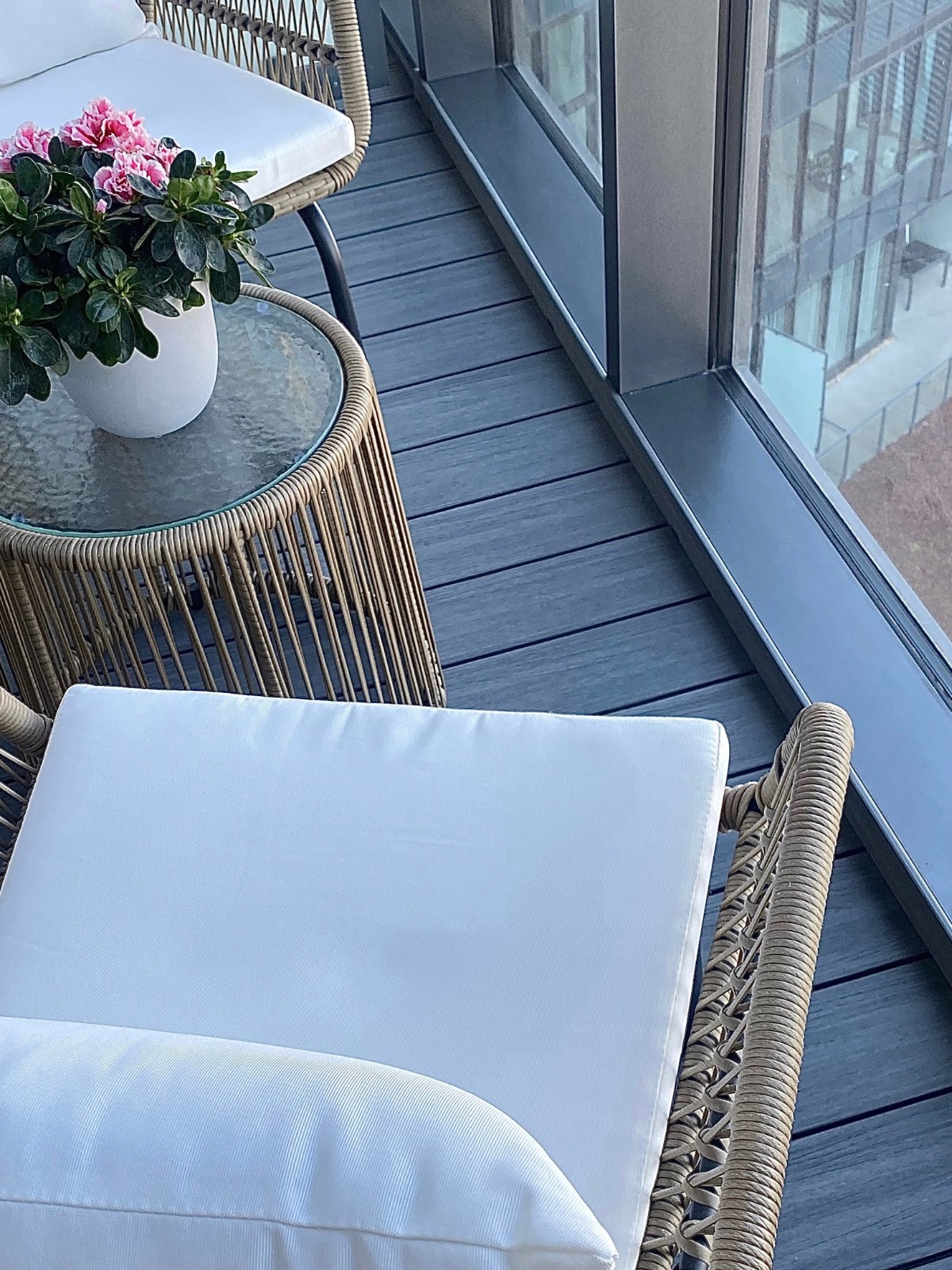 Balkon z rattanowymi meblami i kwiatami w doniczce na szklanym stoliku, widok na miasto przez duże okno. Szare deski podłogowe.