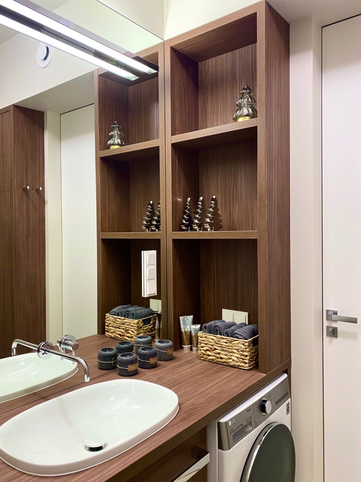 Nowoczesna łazienka z umywalką nablatową, drewnianymi półkami z dekoracjami i pralką w zabudowie. Minimalistyczny design, ciepłe oświetlenie, eleganckie wykończenie.