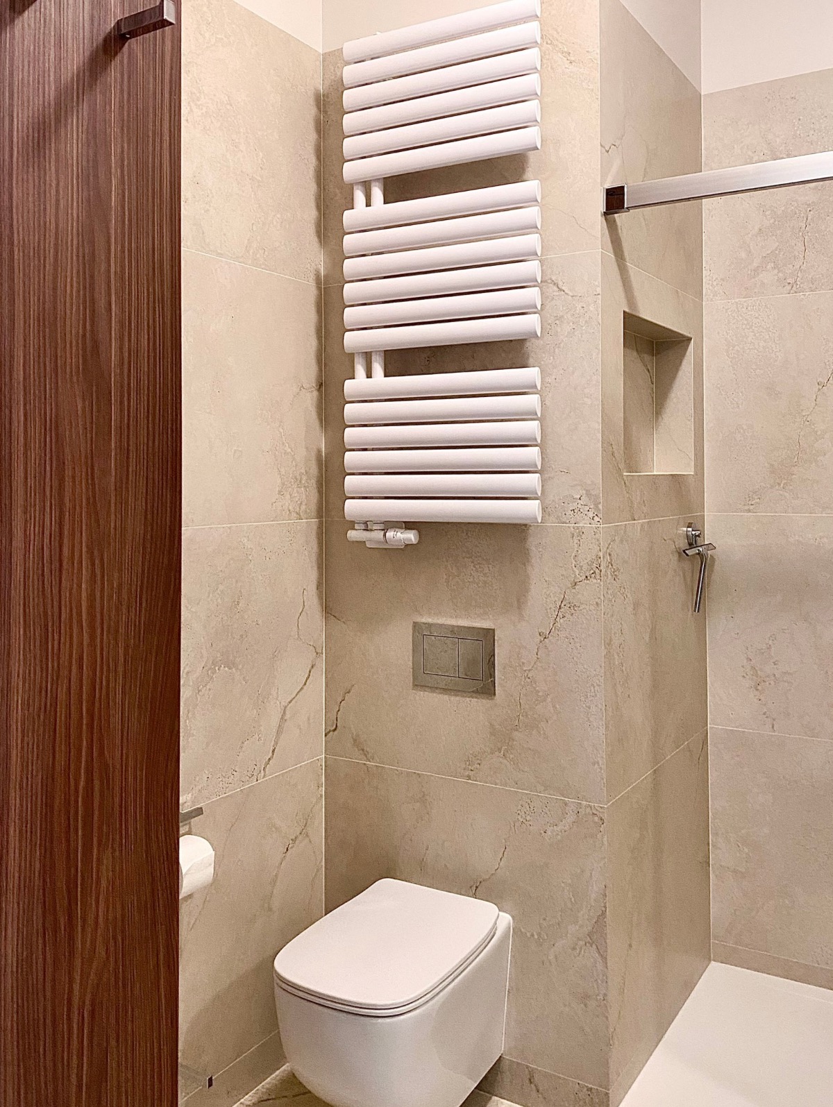 Nowoczesna łazienka z beżowymi płytkami imitującymi marmur, wiszącą toaletą i grzejnikiem drabinkowym. Widoczny fragment kabiny prysznicowej i drewniane drzwi.