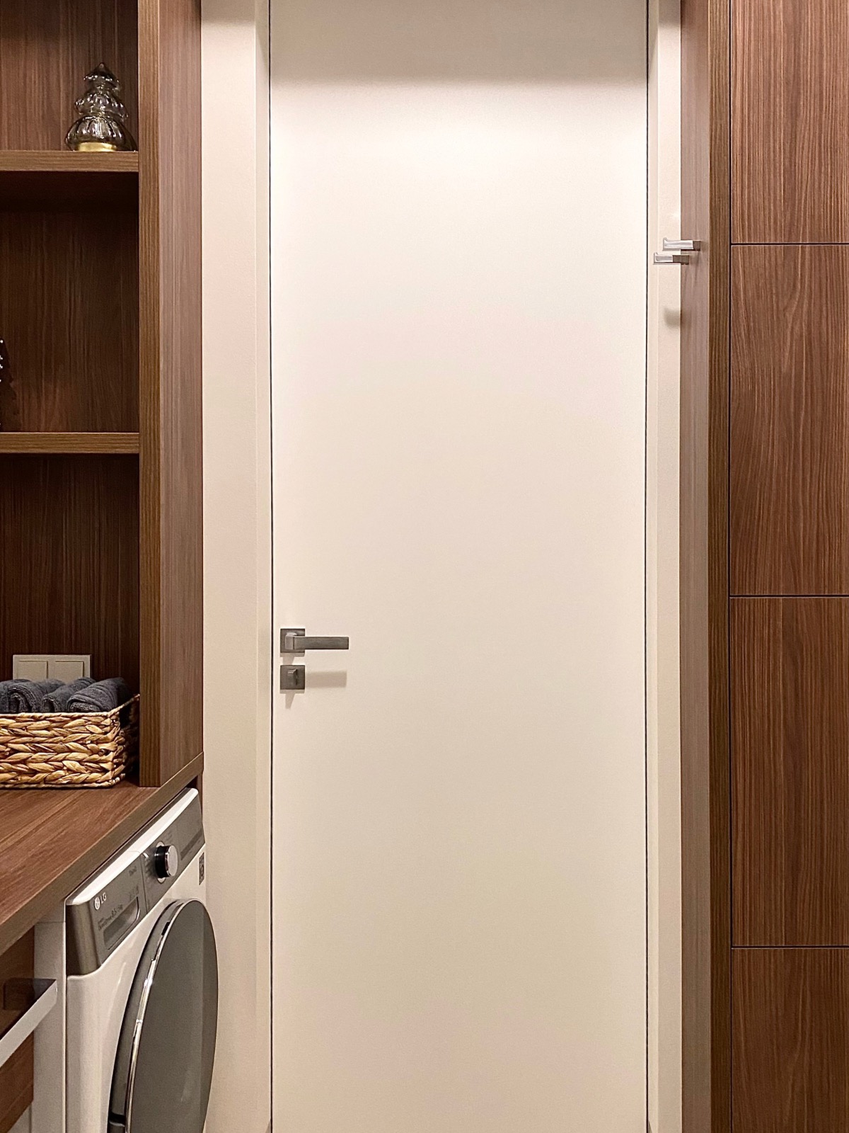 Minimalistyczna aranżacja pralni: białe drzwi z metalową klamką, obok zabudowa z drewnianymi szafkami i pralka. Na półce koszyk z ręcznikami i dekoracja.