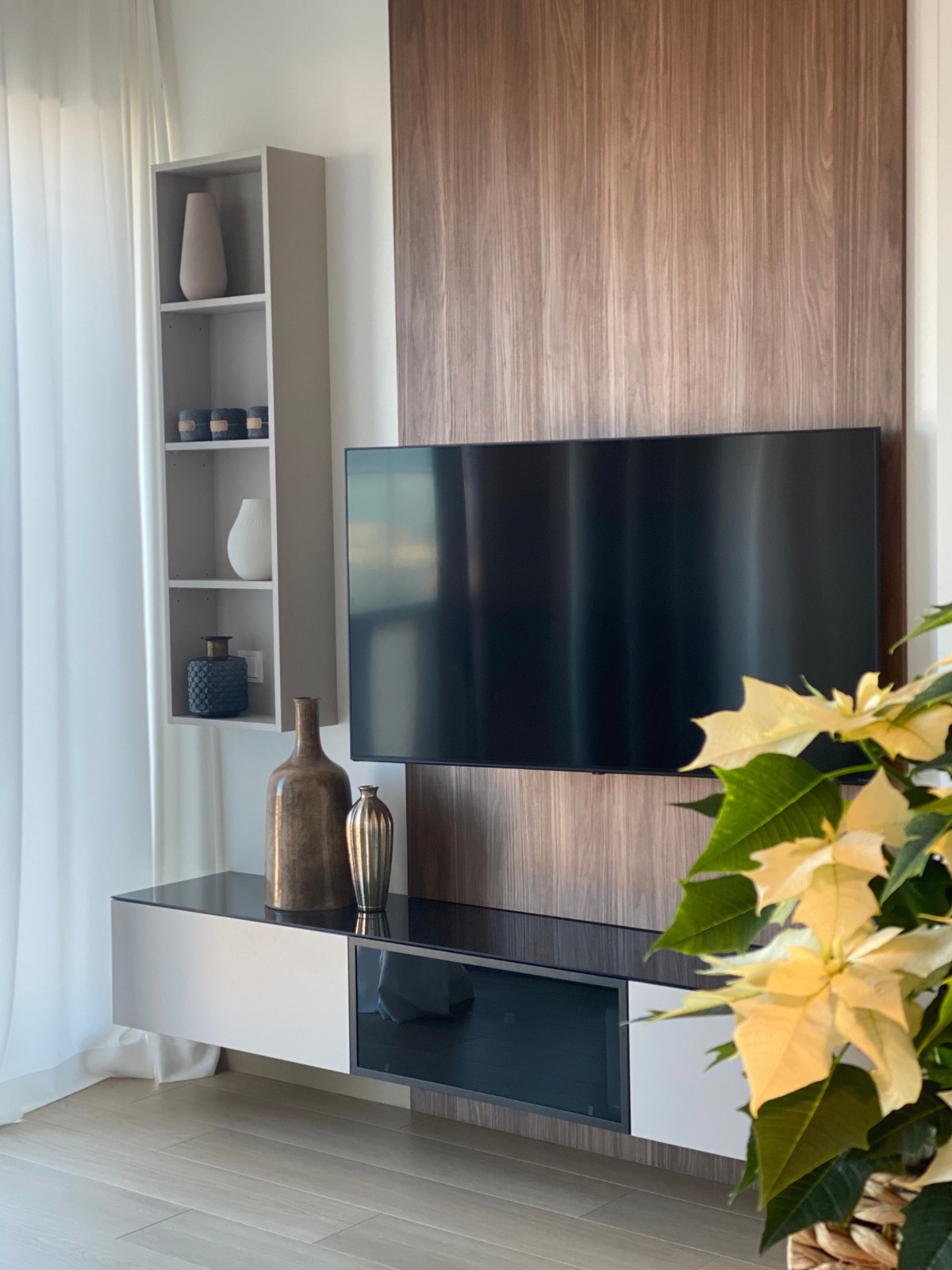 Nowoczesna aranżacja salonu z telewizorem na drewnianym panelu, szafką RTV i regałem z dekoracyjnymi wazonami. Poinsecja w doniczce dodaje akcentu kolorystycznego.