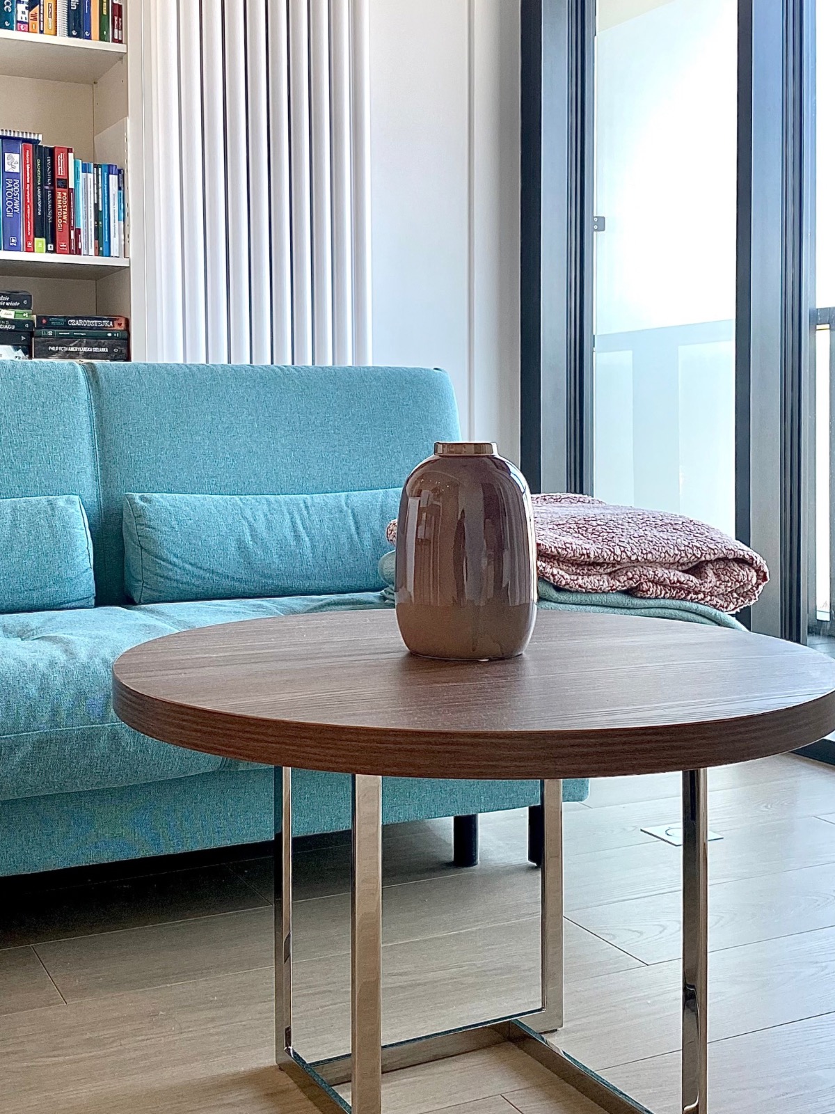 Turkusowa sofa z poduszkami, okrągły stolik kawowy z wazonem, regał z książkami i balkon w tle. Jasne, nowoczesne wnętrze pokoju dziennego.