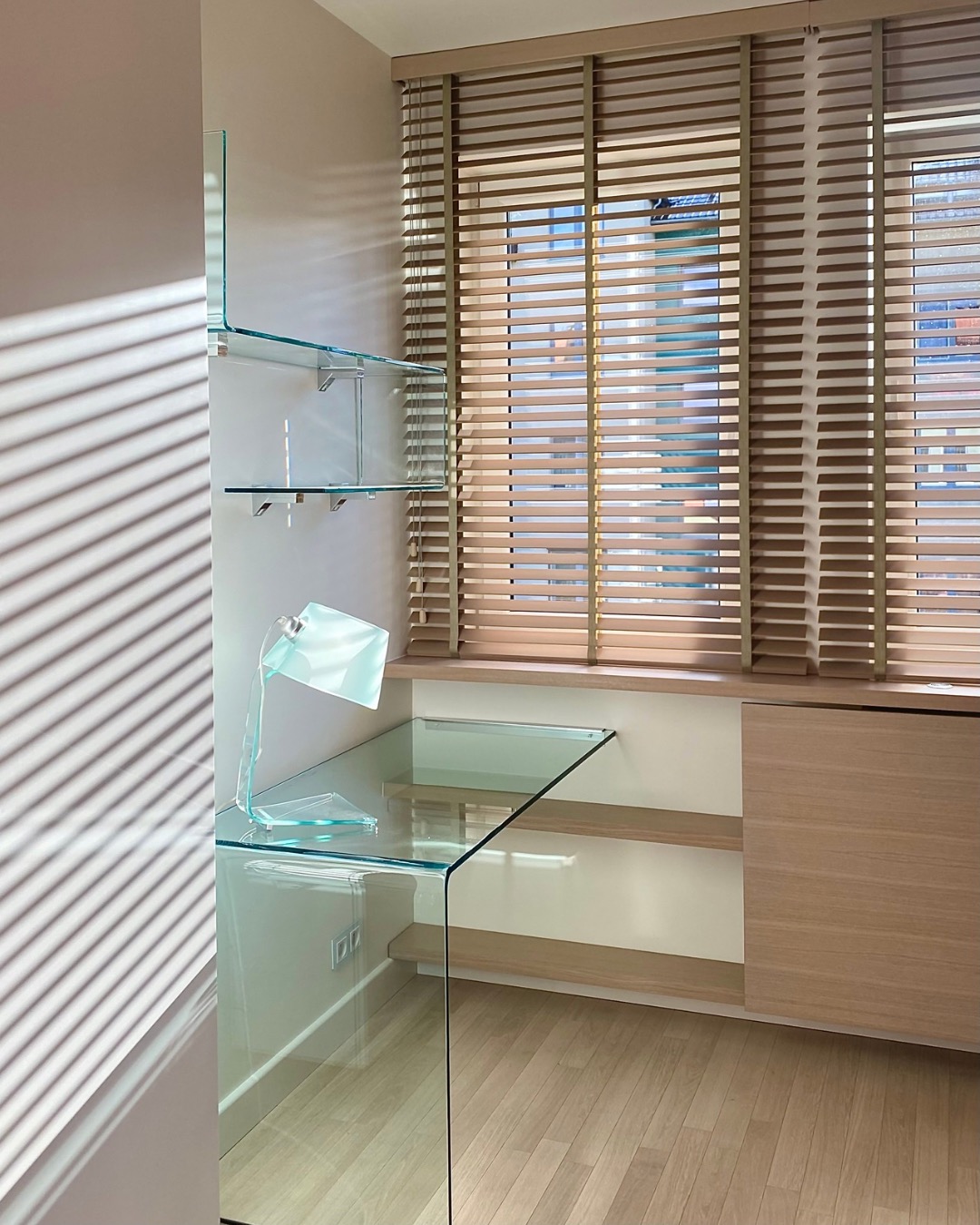 Nowoczesny gabinet z szklanym biurkiem i półkami, drewniane żaluzje w oknie, minimalistyczny design, gra świateł na ścianie, jasne drewno na podłodze.