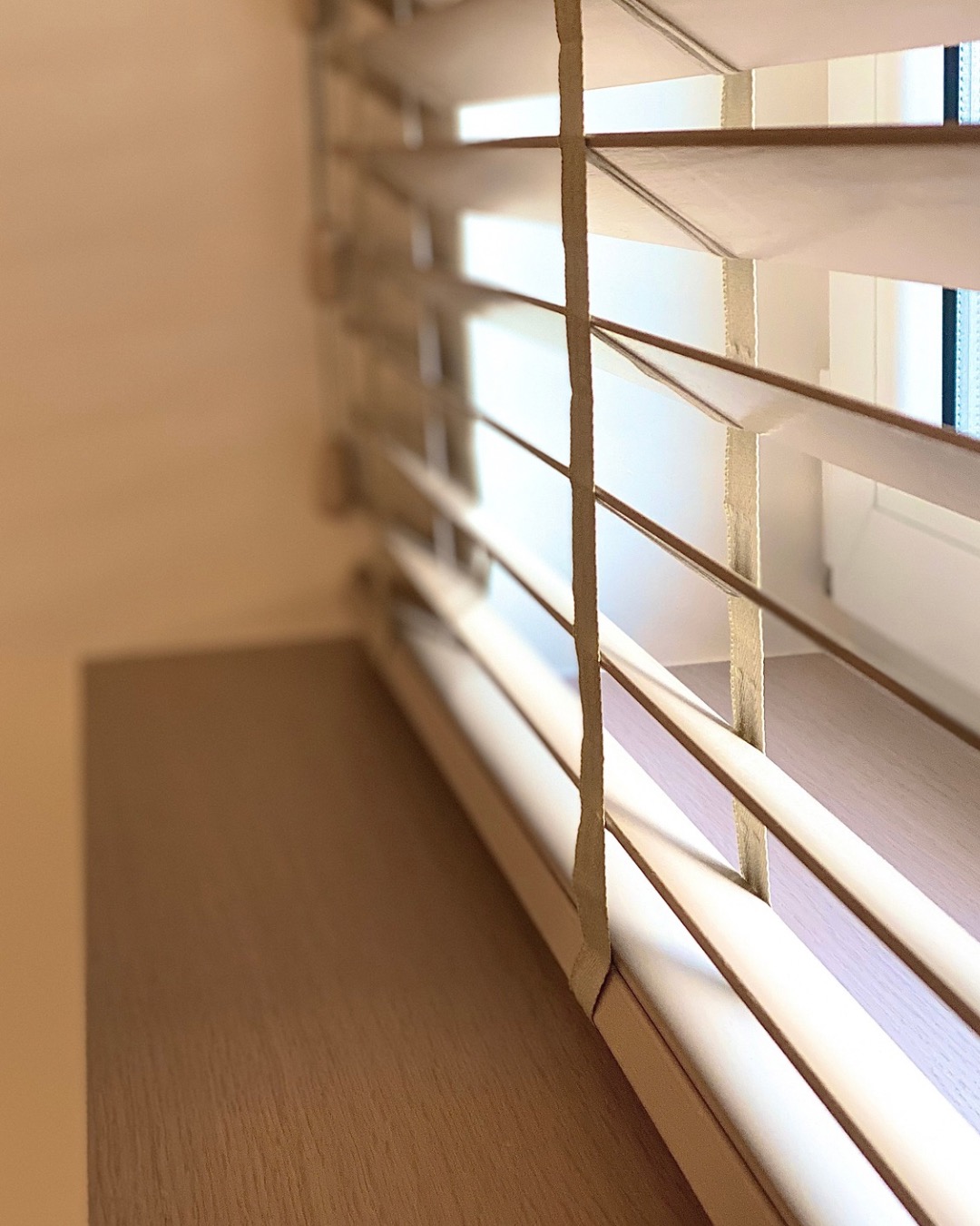 Drewniane żaluzje w ciepłym odcieniu, zamontowane w oknie. Widoczne lamele i sznurki, z delikatnym światłem wpadającym przez szczeliny. Ujęcie z bliska, z perspektywą.