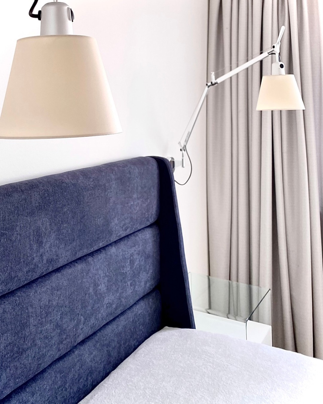 Nowoczesna sypialnia: tapicerowane wezgłowie w odcieniach granatu, lampy z beżowymi abażurami i minimalistyczna szafka nocna z przezroczystego szkła.