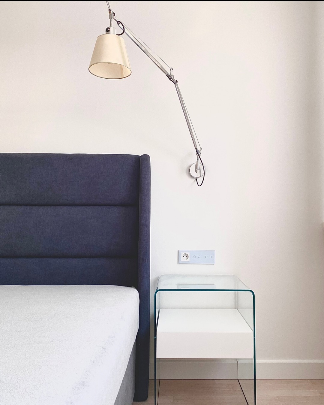 Minimalistyczna sypialnia: łóżko z granatowym zagłówkiem, szklany stolik nocny z białą szufladą, regulowana lampa ścienna i gniazdko z czterema włącznikami.