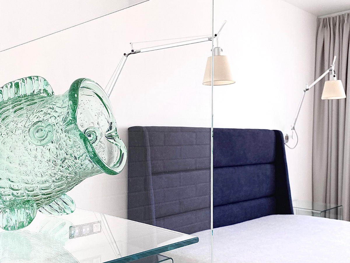 Nowoczesna sypialnia z szarą tapetowaną ścianą za łóżkiem, lampką nocną i szklaną rybą na stoliku. Widok przez szklaną ścianę.