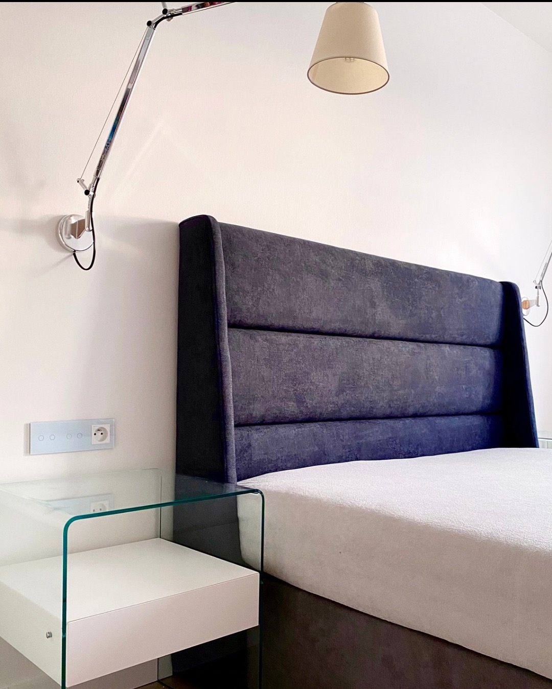 Nowoczesna sypialnia: wezgłowie łóżka z ciemnej tkaniny, szklany stolik nocny z białą półką, lampa ścienna i sufitowa w minimalistycznym stylu.