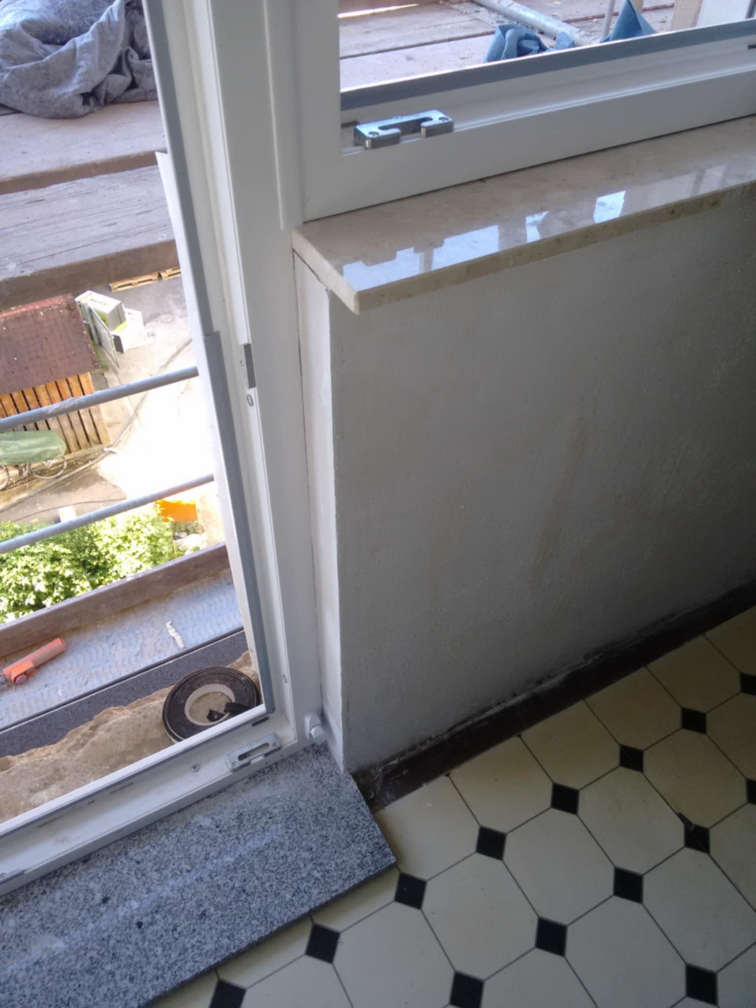 Montaż parapetu z kamienia w oknie. Widoczne nowe, białe okno, szary kamienny parapet wewnętrzny i fragment podłogi z wzorem geometrycznym.