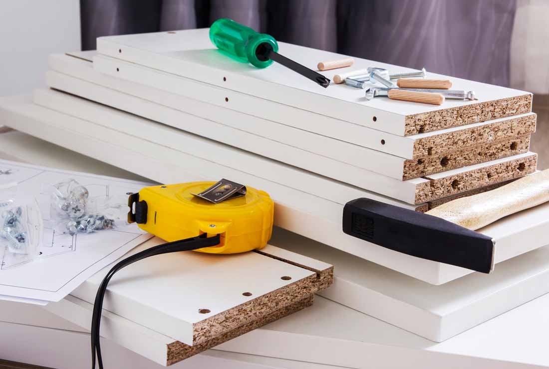 Białe elementy meblowe z narzędziami: śrubokręt, miarka, kołki i instrukcja. Przygotowanie do montażu mebli. Widoczne krawędzie płyty wiórowej.