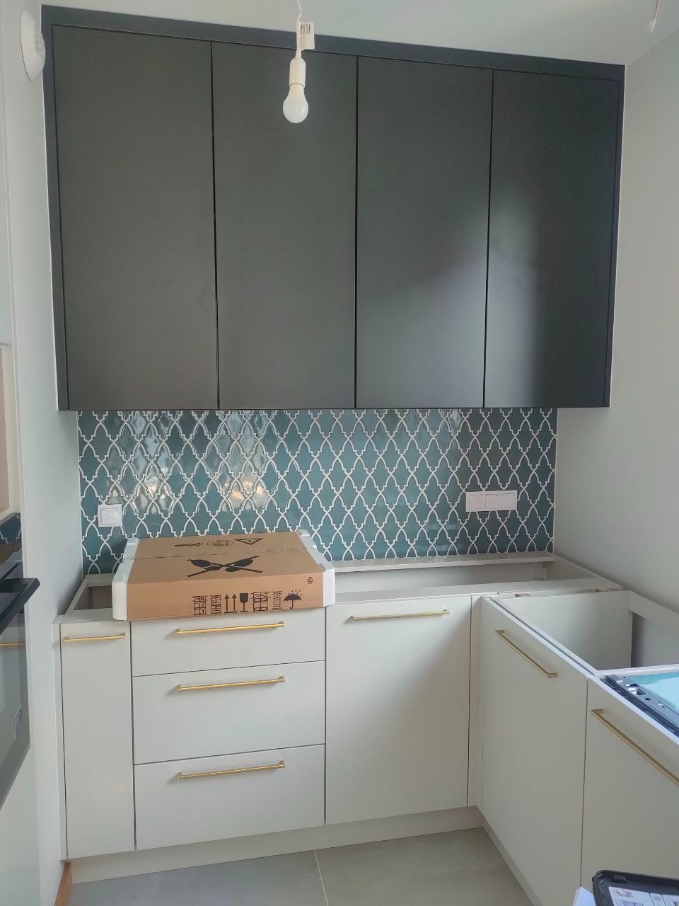 Częściowo zamontowana kuchnia na wymiar: białe szafki z złotymi uchwytami, ciemne szafki górne, wzorzyste płytki nad blatem, karton na blacie.