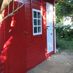 28th Horizon Publishing Serhiy Kravchenko - Czerwony, drewniany domek narzędziowy z białymi akcentami (okno, drzwi) widziany z boku, częściowo przysłonięty gałęziami drzewa. Widoczny fragment chodnika.