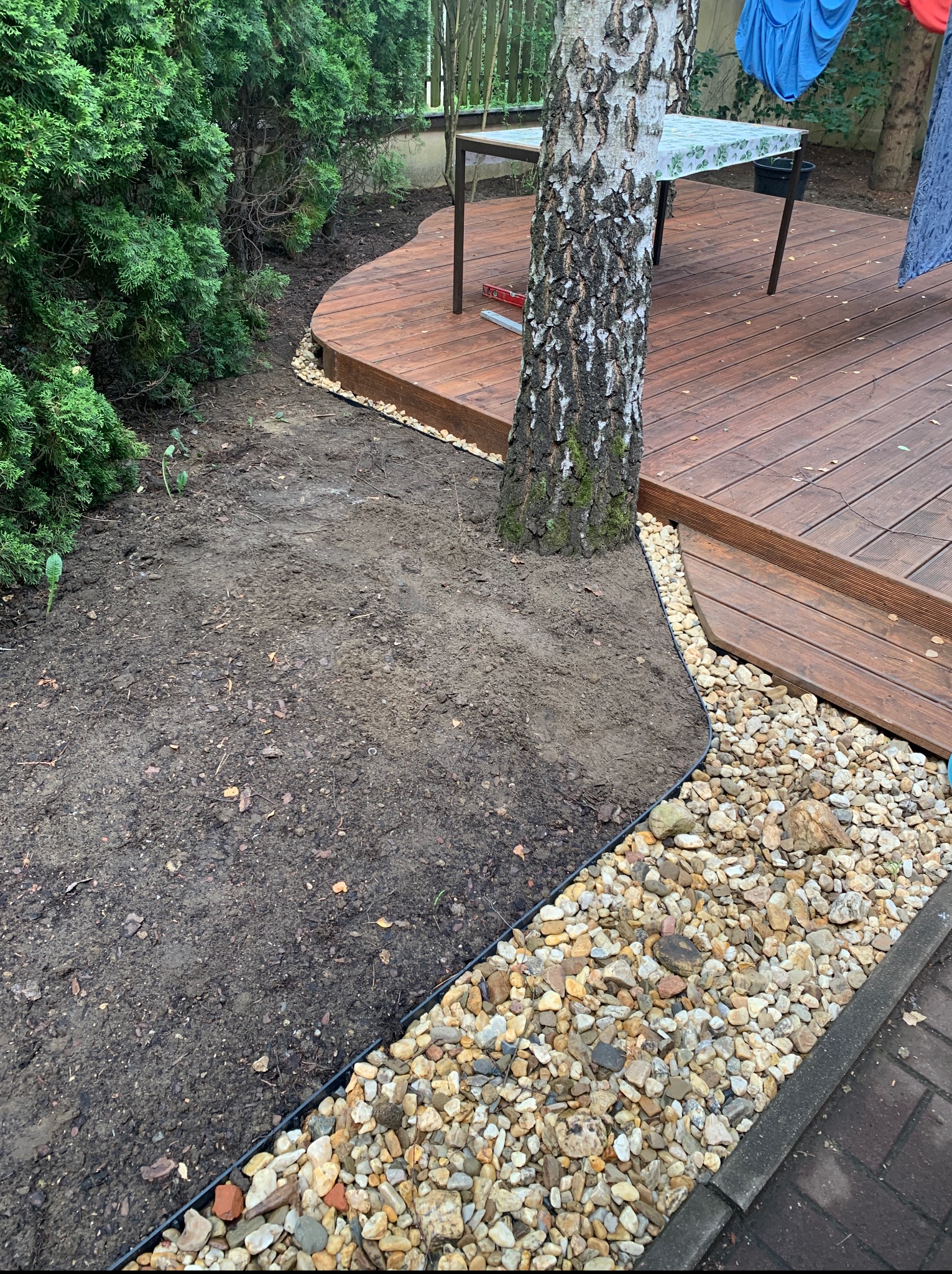 Drewniany taras otoczony rabatą z kamyków i ziemią pod przyszłą zieleń, obok rosną drzewa i krzewy, w tle stół ogrodowy.