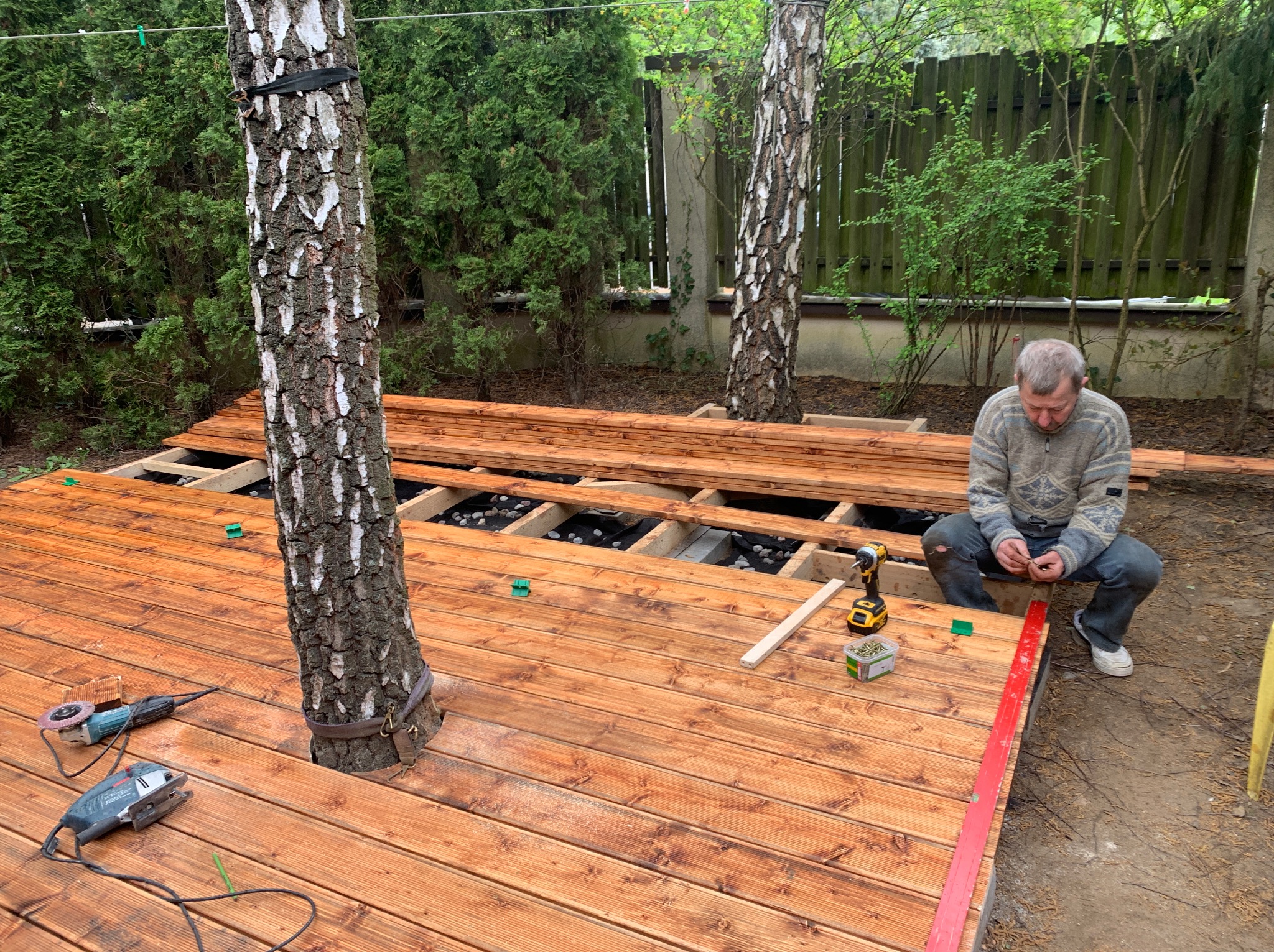 Budowa drewnianego tarasu wokół drzew. Widoczne deski, narzędzia, elementy konstrukcji i pracownik w trakcie montażu na zewnątrz budynku.