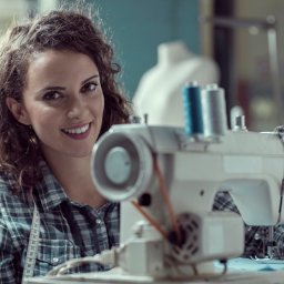 TOPQ BUCIOR BUKOWSKI SPÓŁKA JAWNA - Uśmiechnięta krawcowa przy maszynie do szycia w szwalni, z miarą na szyi. W tle manekin i nici.