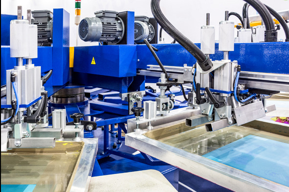 Maszyna do sitodruku z niebieską konstrukcją, srebrnymi elementami i przezroczystymi ramami. Widoczne silniki i przewody. Detal z bliska.