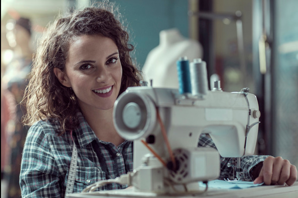 Uśmiechnięta krawcowa przy maszynie do szycia w szwalni, z miarą na szyi. W tle manekin i nici.