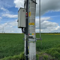 Betonowy słup energetyczny z osprzętem, tabliczkami ostrzegawczymi i skrzynką elektryczną na tle zielonego pola i błękitnego nieba z chmurami.