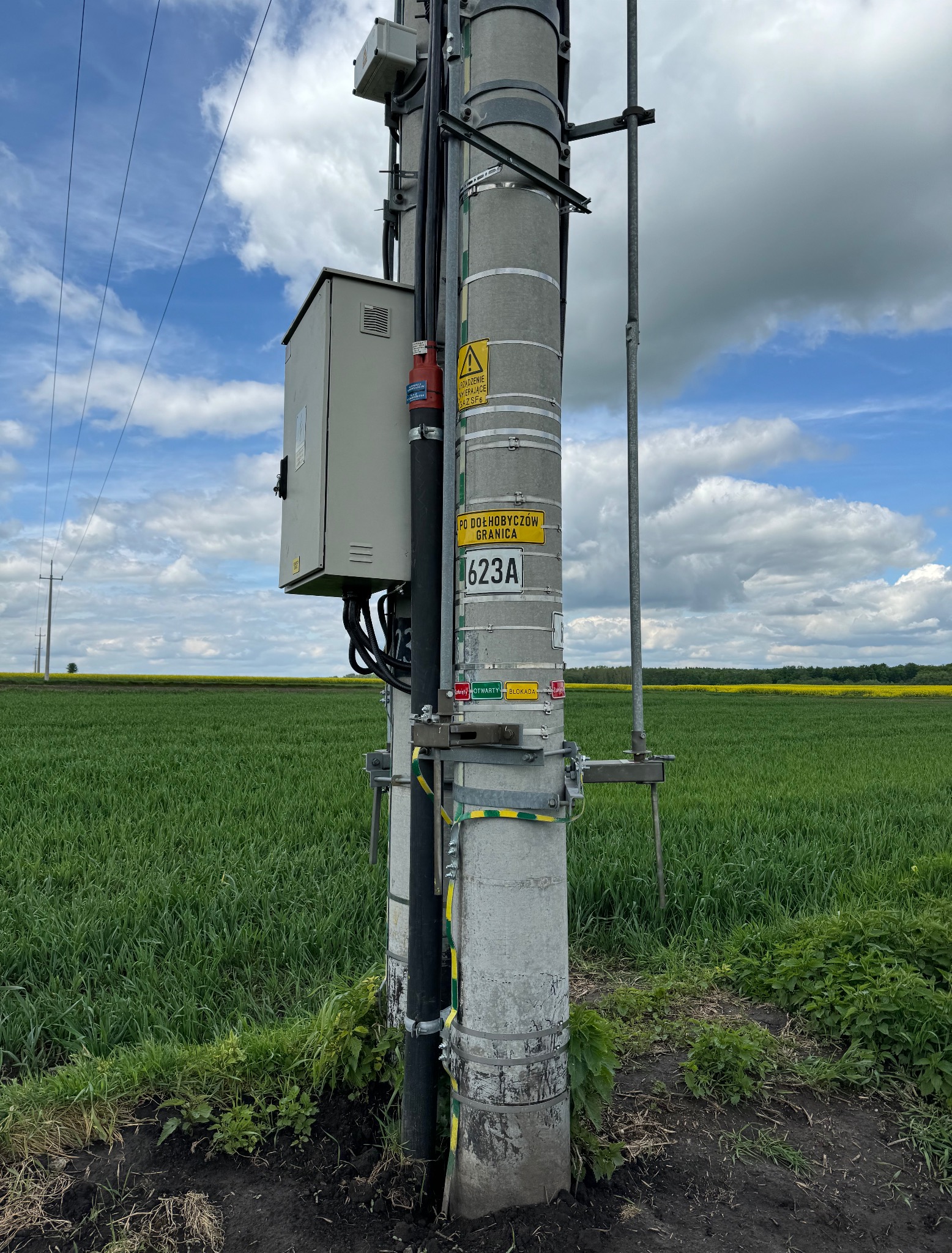 Betonowy słup energetyczny z osprzętem, tabliczkami ostrzegawczymi i skrzynką elektryczną na tle zielonego pola i błękitnego nieba z chmurami.