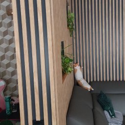 Piotr Wiśniewski - Nowoczesna aranżacja wnętrza z drewnianymi lamelami w odcieniach beżu i szarości. Dekoracyjne rośliny i poduszki dopełniają wystrój salonu z szarą sofą.