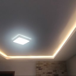 Piotr Wiśniewski - Podwieszany sufit z oświetleniem LED wzdłuż krawędzi i centralną lampą kwadratową, fragment ściany z cegły dekoracyjnej w dolnej części kadru.