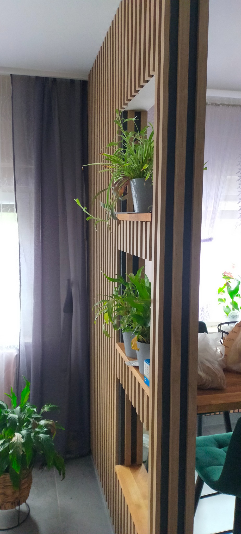 Drewniana ścianka działowa z lamelami i półkami, na których stoją doniczki z roślinami. Widoczne szare zasłony i fragment wnętrza z zielonym krzesłem.