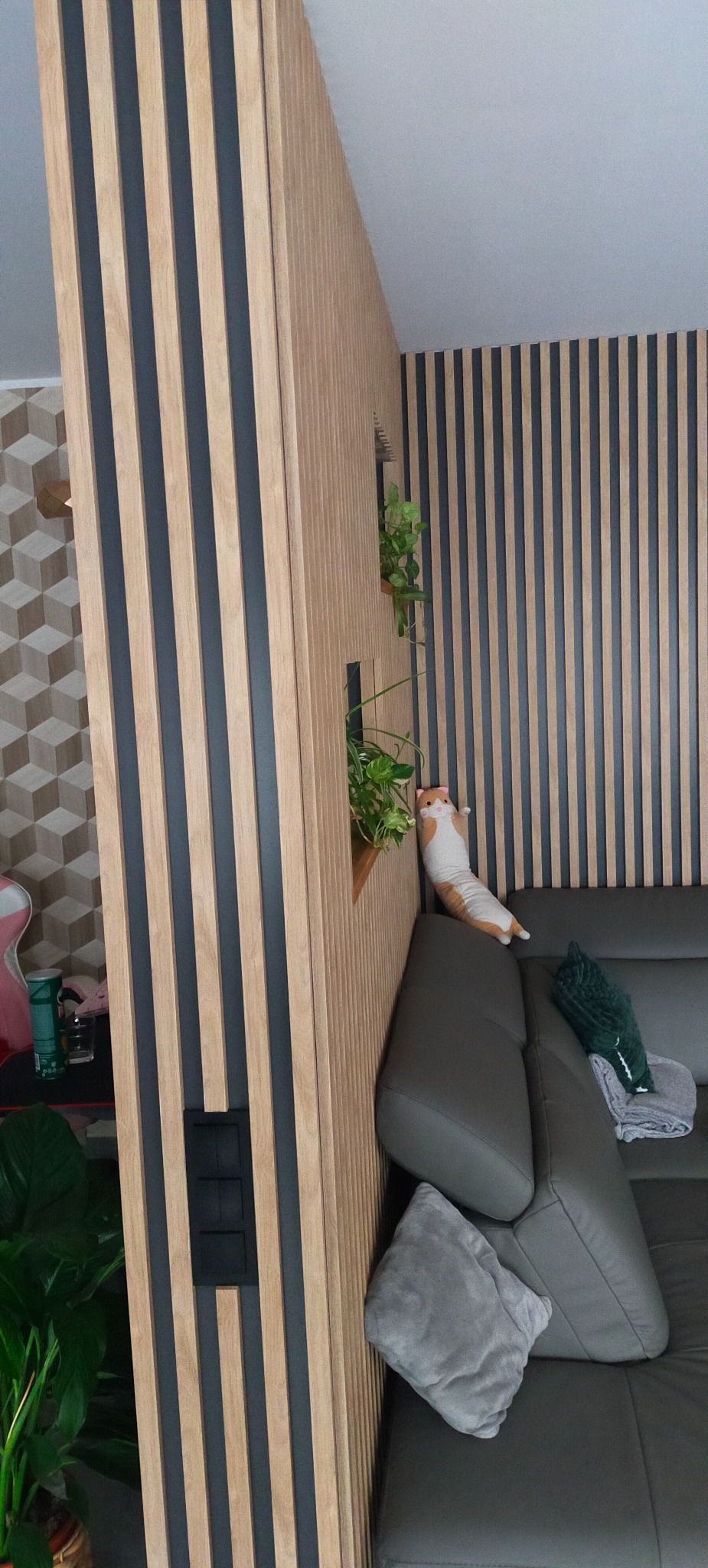 Nowoczesna aranżacja wnętrza z drewnianymi lamelami w odcieniach beżu i szarości. Dekoracyjne rośliny i poduszki dopełniają wystrój salonu z szarą sofą.