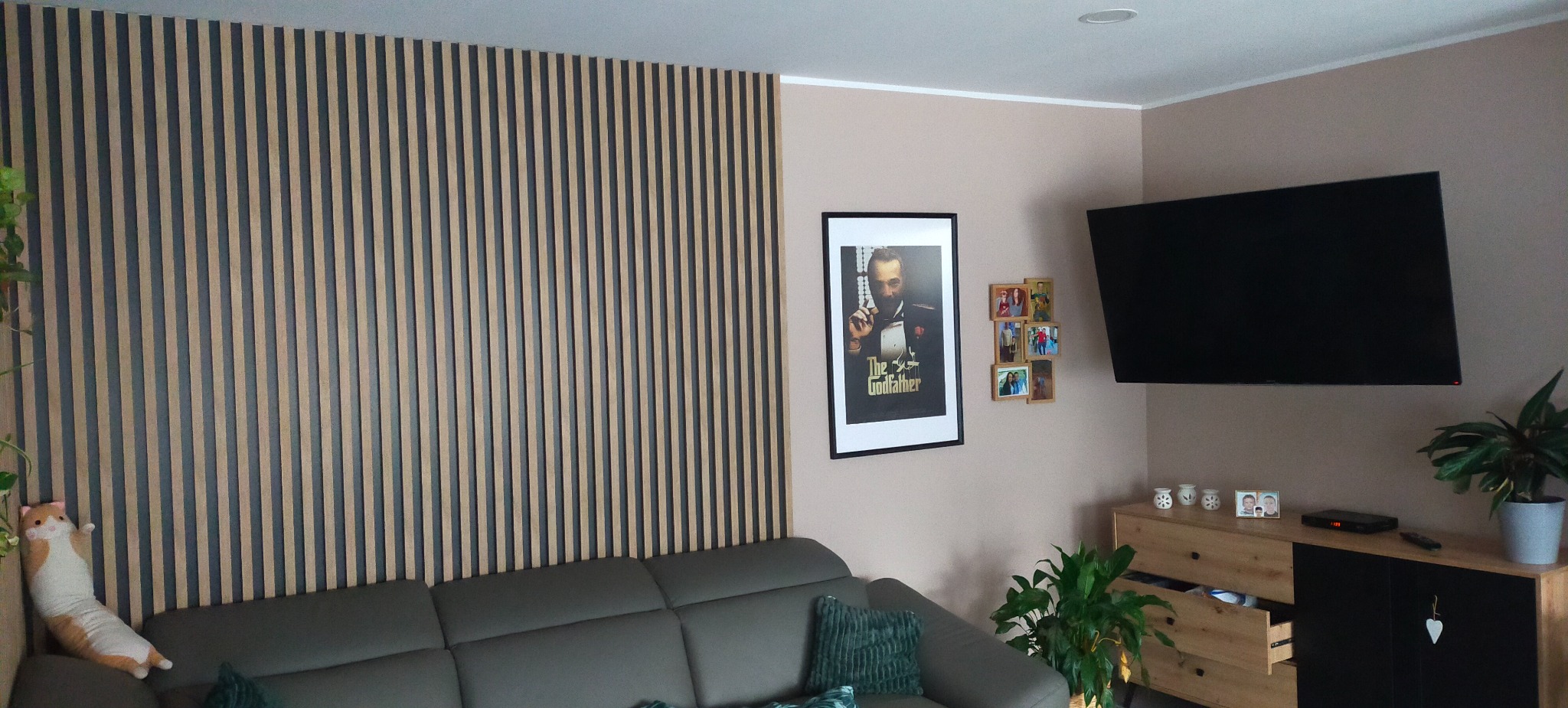 Salon z panelami dekoracyjnymi na ścianie za szarą kanapą. Na ścianie wisi telewizor i plakat. Komoda z otwartą szufladą i kwiaty doniczkowe dopełniają aranżację.