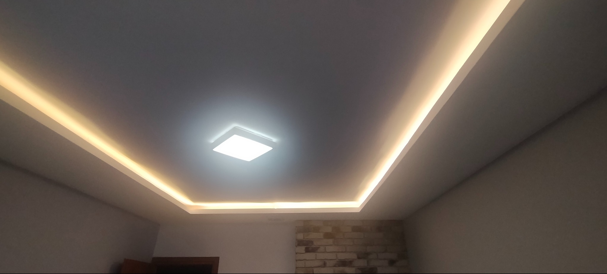 Podwieszany sufit z oświetleniem LED wzdłuż krawędzi i centralną lampą kwadratową, fragment ściany z cegły dekoracyjnej w dolnej części kadru.