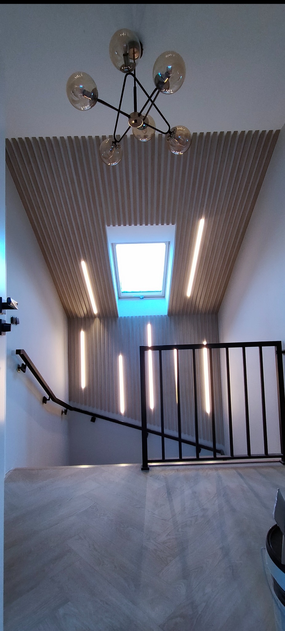 Nowoczesne wykończenie klatki schodowej z drewnianymi lamelami na suficie, oknem dachowym i podświetleniem LED. Minimalistyczna balustrada.