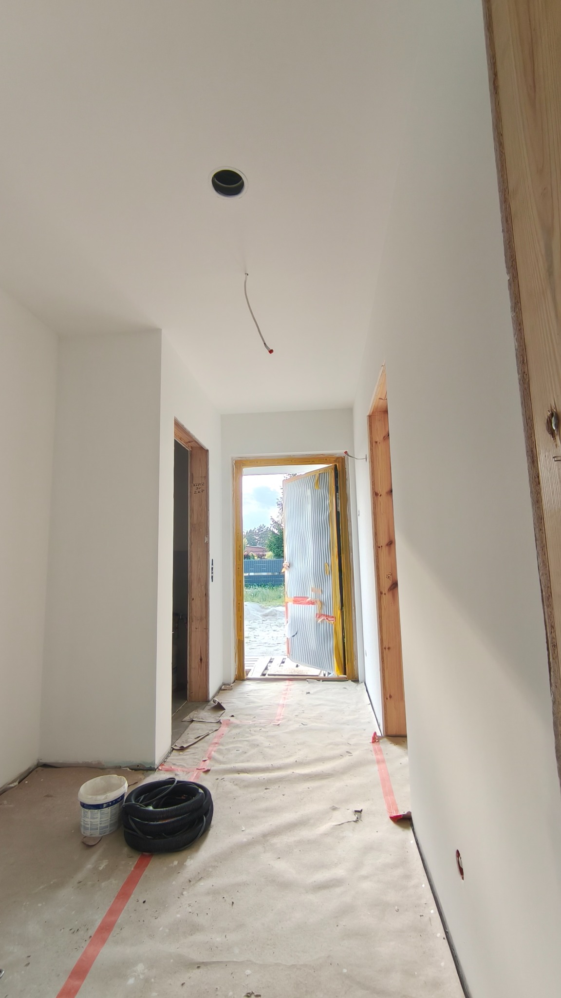 Białe, świeżo pomalowane ściany w korytarzu, zabezpieczona podłoga, widoczne kable i puszka w suficie, surowe ościeżnice drzwi. Prace wykończeniowe w nowym domu.