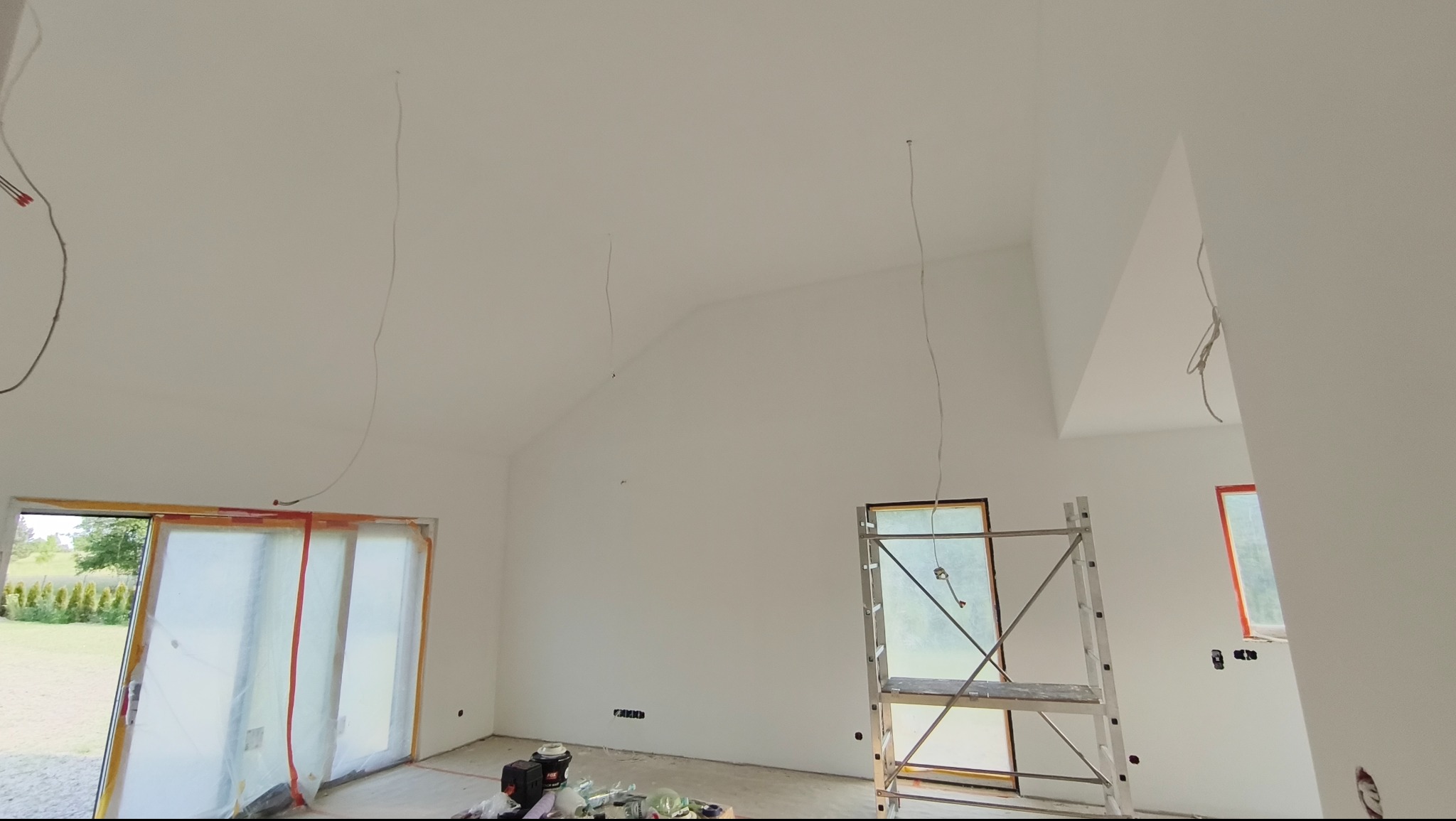 Białe wnętrze w trakcie odświeżania, widoczne kable zwisające z sufitu, okna zabezpieczone folią i rusztowanie. Ściany i sufit świeżo pomalowane na biało.