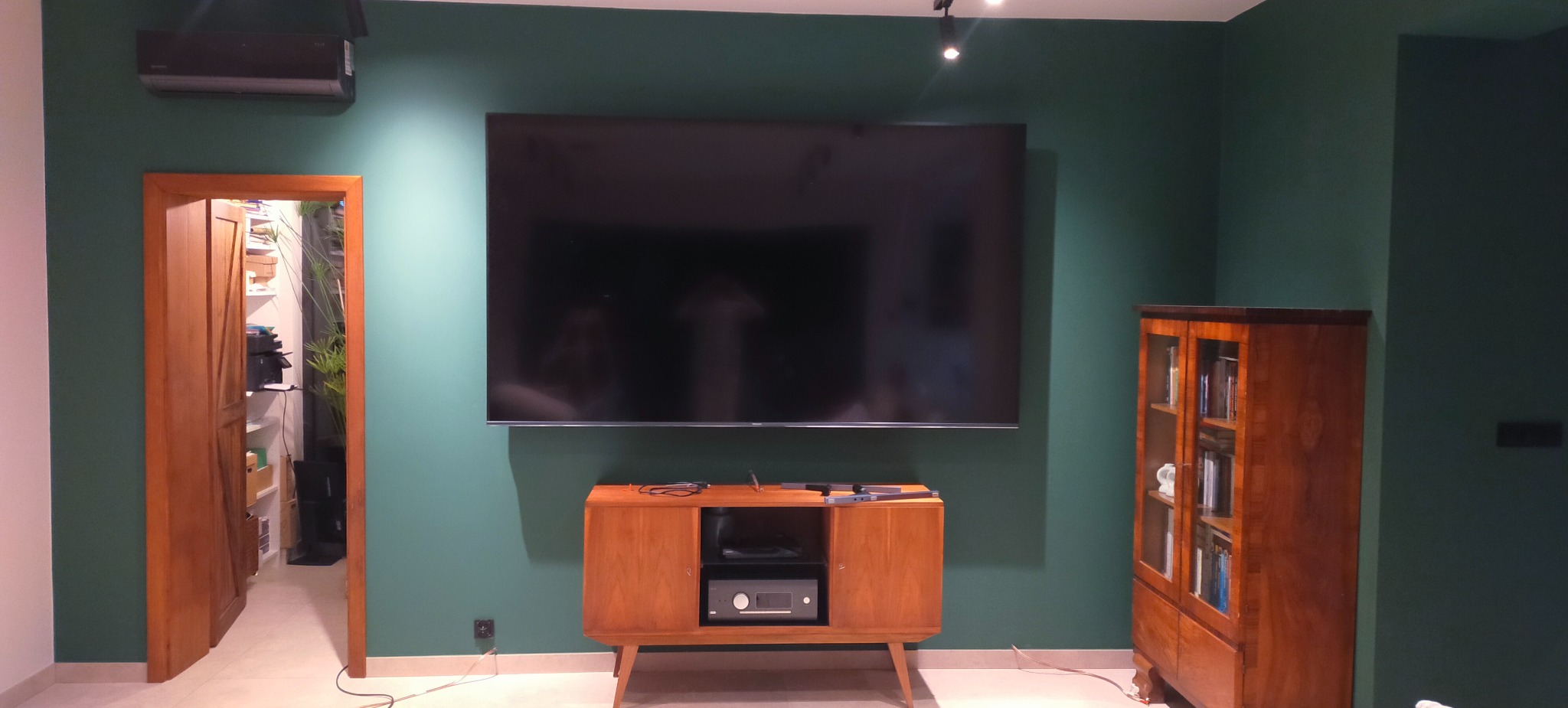 Salon po malowaniu ścian na ciemnozielono. Telewizor na ścianie, drewniana szafka RTV i regał. Drzwi otwarte do jasnego pomieszczenia. Oświetlenie punktowe.