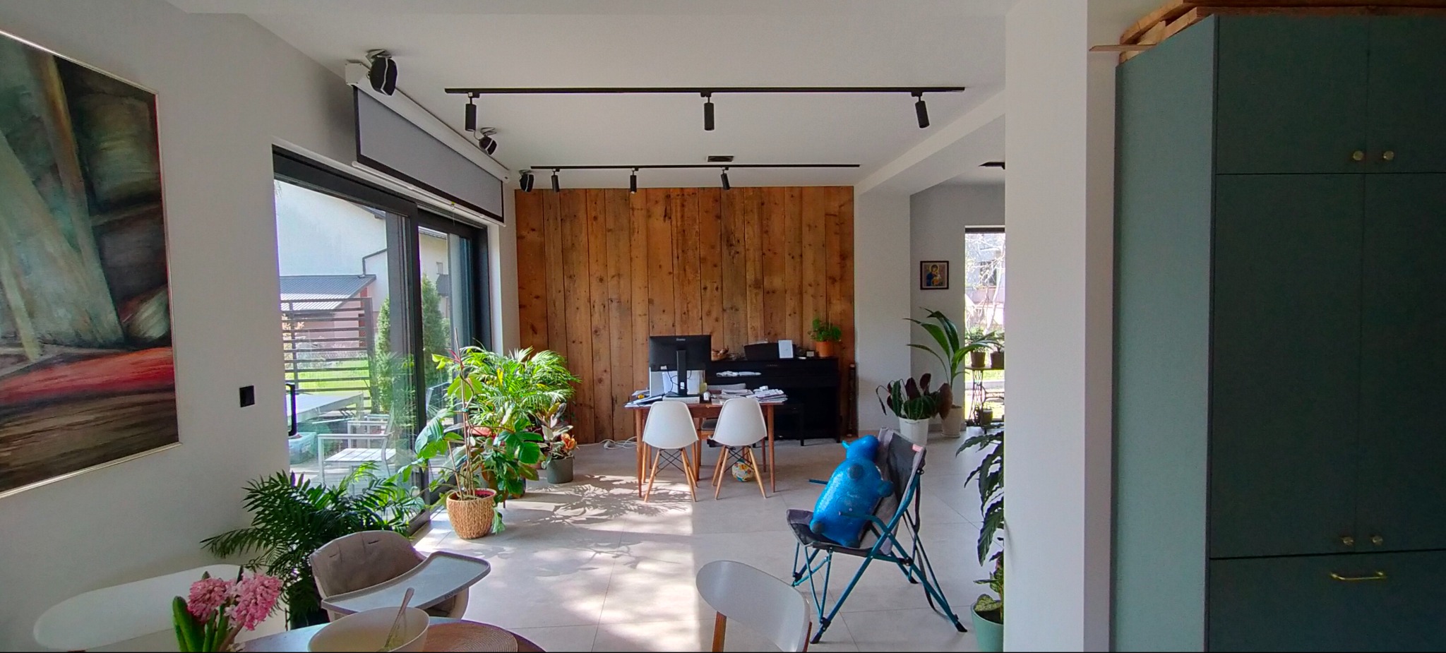 Jasne wnętrze z drewnianą ścianą, oświetleniem szynowym i roślinami doniczkowymi. Widoczny fragment obrazu i zabudowa meblowa w odcieniach zieleni.