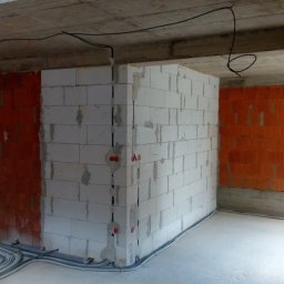 Surowy stan: kable elektryczne w peszlach ułożone wzdłuż ścian z białego i czerwonego bloczka, gniazdka w ścianach, przewody pod sufitem.