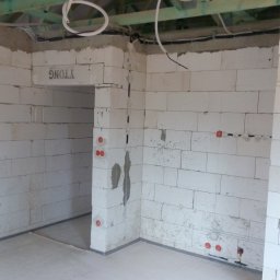 Wnętrze w trakcie budowy z białymi ścianami z betonu komórkowego. Widoczne przewody elektryczne w ścianach i pod sufitem. Surowy stan, brak wykończenia.