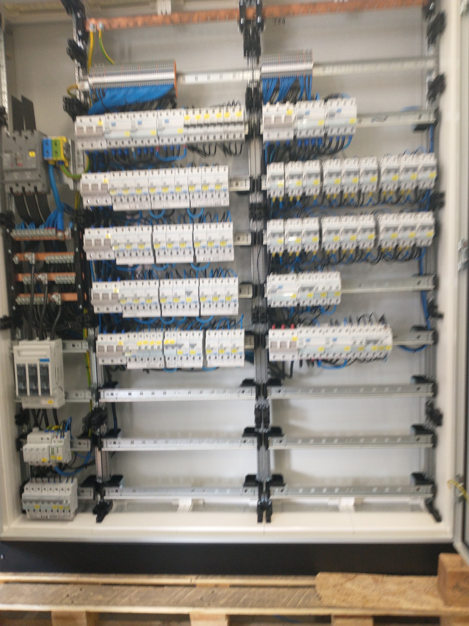 Rozdzielnica elektryczna z licznymi bezpiecznikami, przewodami i szynami, zamontowana wewnątrz białej obudowy. Widoczne szczegóły instalacji elektrycznej.