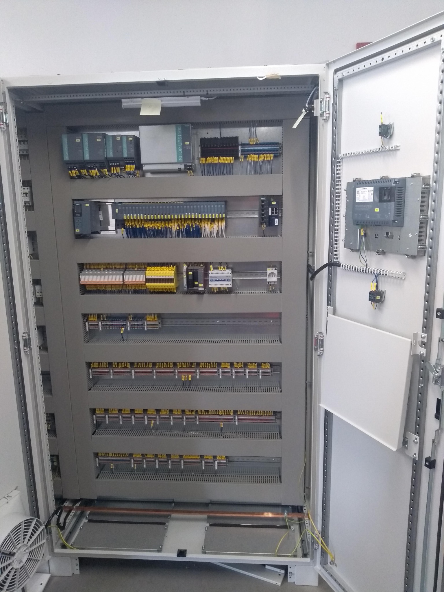 Otwarta szafa sterownicza z modułami Siemens, widoczne liczne przewody i złącza. Elementy automatyki przemysłowej w szarej obudowie.