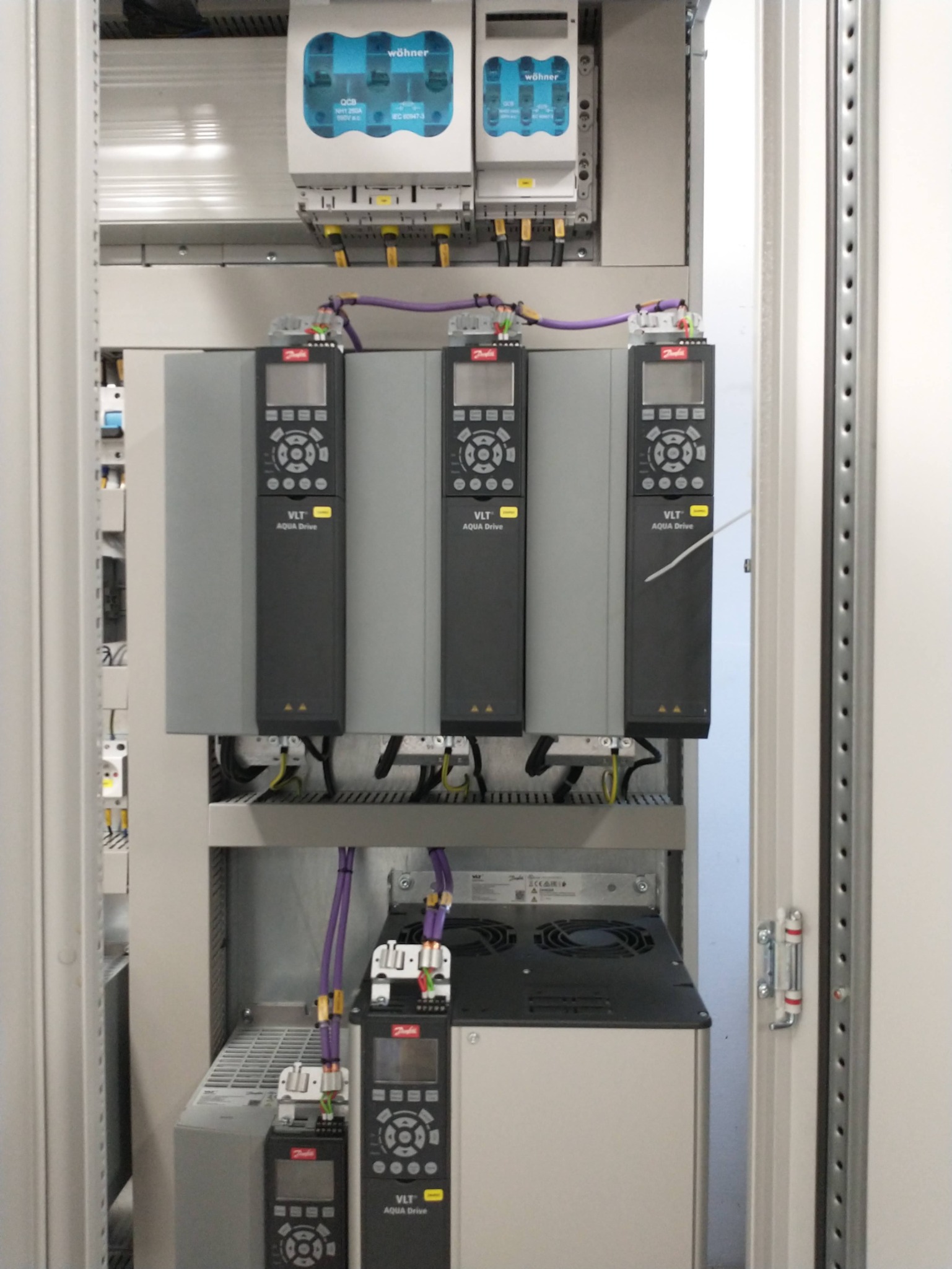Rozdzielnica elektryczna z zamontowanymi urządzeniami Danfoss VLT Aqua Drive, widoczne kable i zabezpieczenia Wöhner. Całość w metalowej szafie.