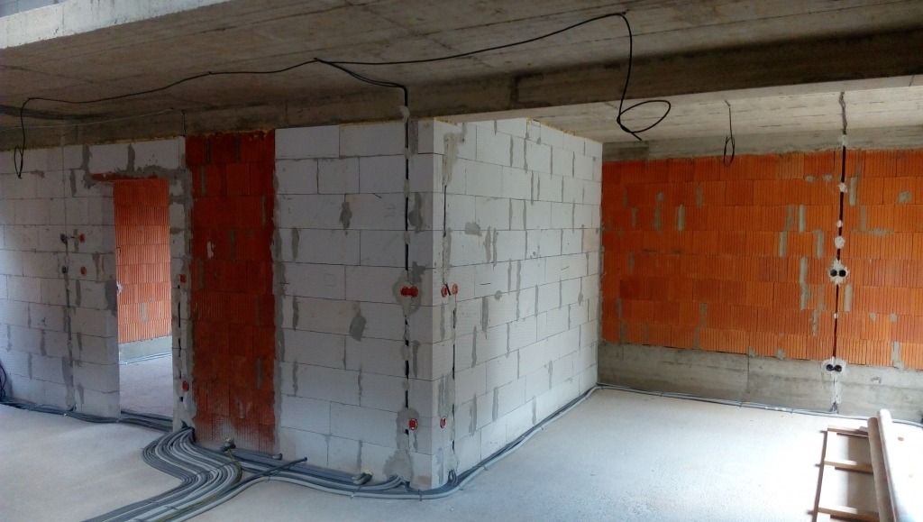 Surowy stan: kable elektryczne w peszlach ułożone wzdłuż ścian z białego i czerwonego bloczka, gniazdka w ścianach, przewody pod sufitem.