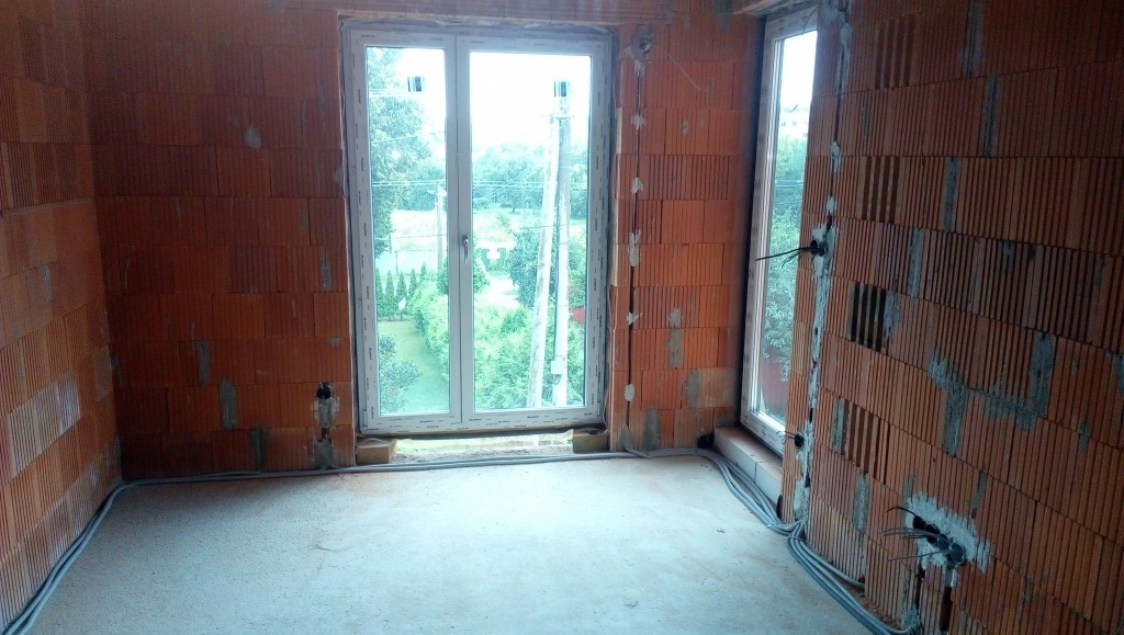 Surowy pokój z czerwonej cegły dziurawki. Widoczne kable elektryczne wzdłuż ścian i pionowe bruzdy w murze pod instalacje. Dwa okna z widokiem na zieleń.