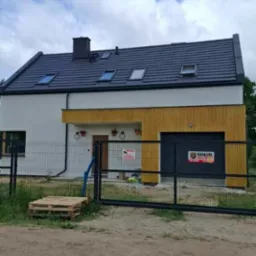 Nowoczesny dom z ocieploną elewacją w kolorach bieli i drewna, widoczny zza metalowego ogrodzenia. Dach pokryty czarną dachówką, okna dachowe. Paleta przed ogrodzeniem.