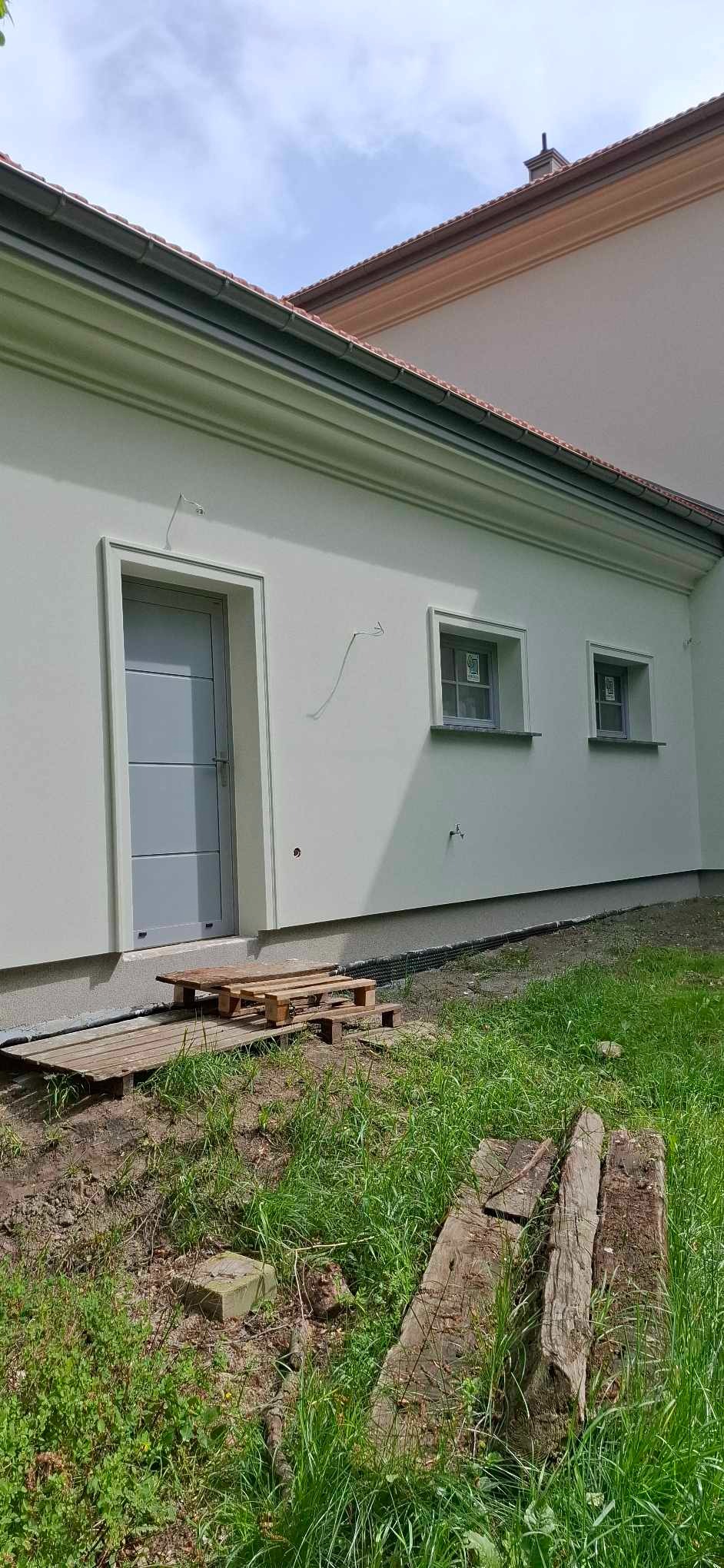 Jasna, otynkowana elewacja domu z szarą stolarką okienną i drzwiową. Widoczne palety i drewniane elementy na trawniku przed budynkiem.