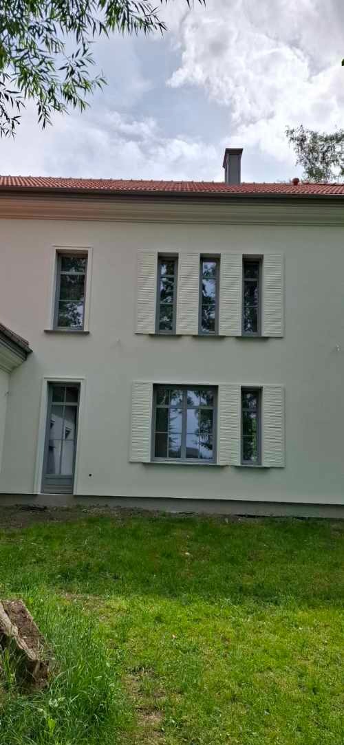 Biała, dwukondygnacyjna elewacja domu z szarymi oknami i okiennicami. Widoczny fragment dachu z kominem, zielony trawnik i drzewa w tle.